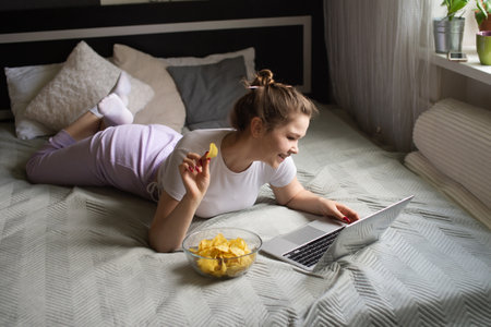 A cute girl is lying on the bed in home clothes eating chips and looking at a laptop