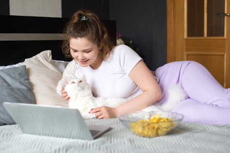 A cute girl is lying on the bed stroking the cat and looking at her laptop, chips in a plateの写真素材