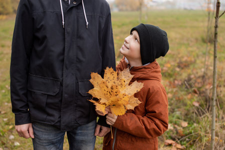 The boy holds a bouquet of autumn leaves and looks at his dad by the handの写真素材