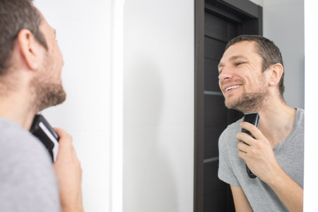 A man shaves his beard with a trimmer , looks in the mirror and smilesの写真素材