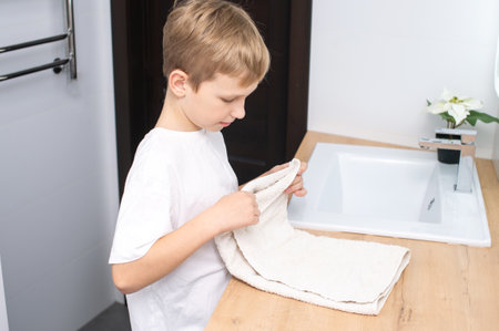 The boy is standing in the bathroom near the washbasin and folds a towelの写真素材