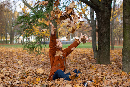 The boy throws up autumn yellow leaves and looks up sitting on the groundの写真素材