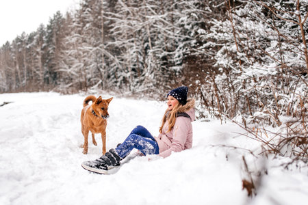 A cute girl is sitting in the snow next to a red dog in the forestの写真素材