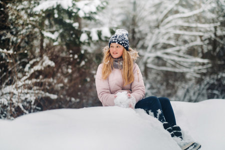 Cute girl with long hair is sitting on a mountain of snow in a hat and making snowballsの写真素材