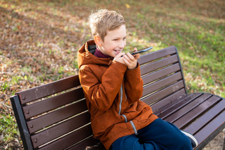 Cute boy records a voice message and laughs while sitting on a park bench in autumnの写真素材
