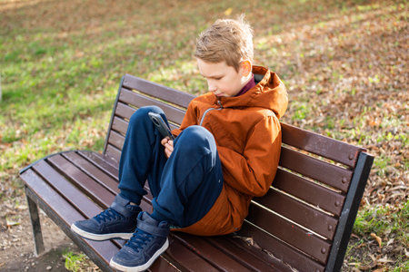 The boy is playing on the phone sitting on a park bench in autumn and smiling. Feet on the benchの写真素材
