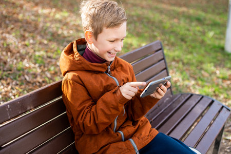 The boy is playing on the phone sitting on a park bench in autumn and smilingの写真素材