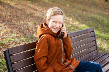 A boy is talking on the phone and laughing while sitting on a park bench in autumnの写真素材