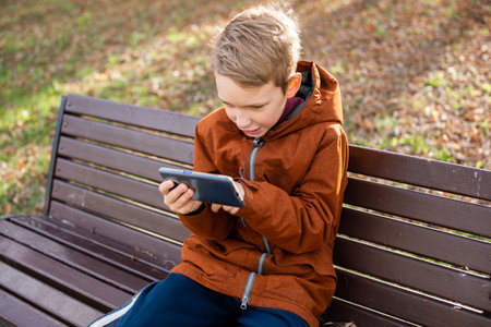 An emotional boy is talking on the phone holding his head and laughing while sitting on a park bench in autumnの写真素材