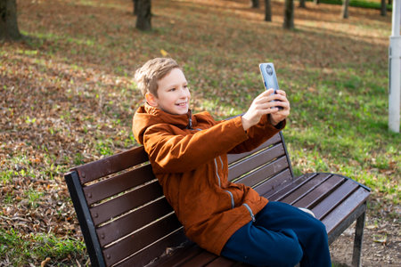 A boy takes a selfie on his phone while sitting on a park bench in autumnの写真素材