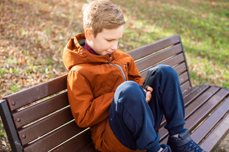 A boy plays on his phone while sitting on a park bench in autumnの写真素材