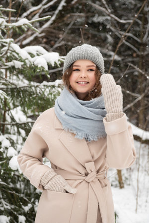 A cute girl is standing under the snow and smiling in the forest in mittens and coatsの写真素材