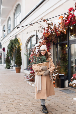 A stylish young woman wearing a hat with branches of nobilis in a craft bag walks around the Christmas cityの写真素材
