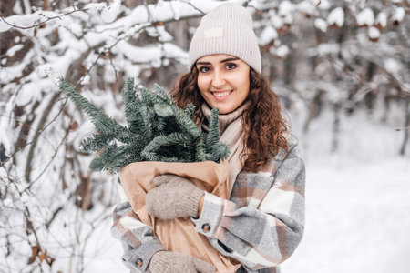 A stylish girl in mittens in a snowy forest holds a kraft bag from a branch of a nobilis fir and smilesの写真素材