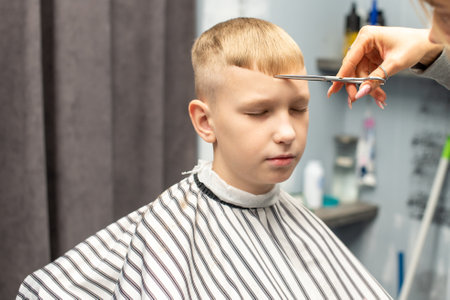 A schoolboy sits with his eyes closed in a barbershop, doing his hair with scissors for haircutsの写真素材