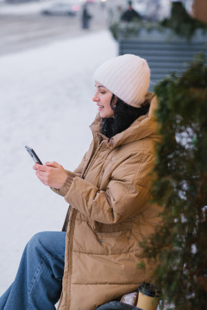 A young girl in a white hat and coat sits on a bench in winter and talks on the phoneの写真素材