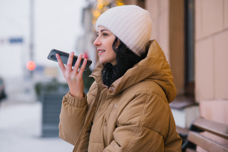A young girl in a hat and coat records a voice message outside in winter and laughsの写真素材