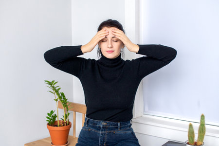 A woman has a headache, sits on a chair and holds her head in her handsの写真素材