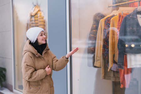 A stylish young woman in a white hat and coat points her hand at the window of a clothing storeの写真素材