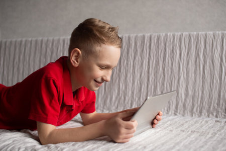 A cute boy in a red T-shirt is lying and playing on a tablet on the couch and laughingの写真素材