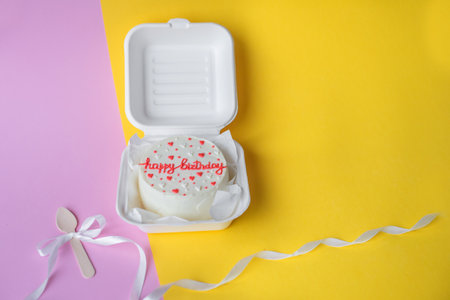 A small bento birthday cake in a box. Wooden spoon with white ribbon. Yellow and pink backgroundの写真素材