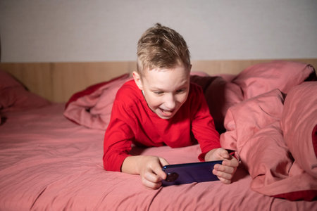 Cute boy woke up and is playing on the phone lying on the bed under the blanketの写真素材