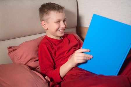 A happy boy in red pajamas is lying in bed reading a book and laughingの写真素材