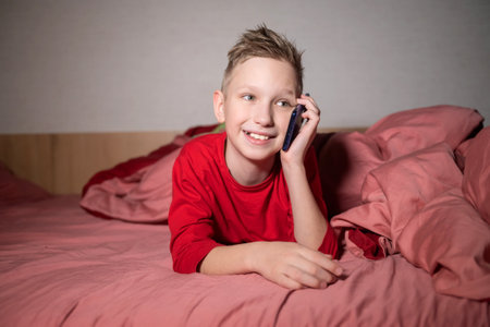 A cute boy has woken up and is calling on the phone lying on the bed under a blanketの写真素材