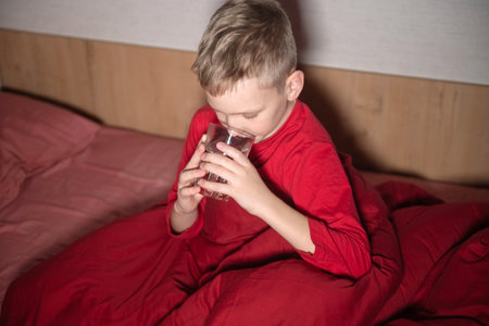 A happy boy is sitting in red pajamas under a blanket and drinking water from a glassの写真素材
