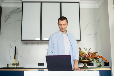 A young man standing in the kitchen is working on a laptop and holding a disposable cupの写真素材