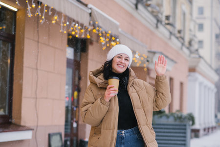 A happy woman walks through the city in winter with a cup of coffee and waves her handの写真素材