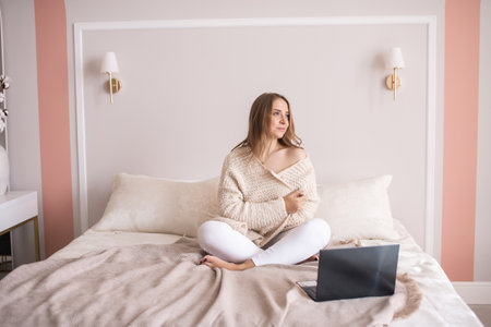 A sad girl in home clothes is sitting on a bed with a laptop and warming herself in a sweaterの写真素材