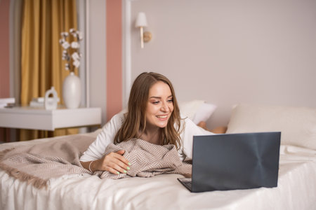 Cute girl is working on a laptop while lying at home on a bedの写真素材