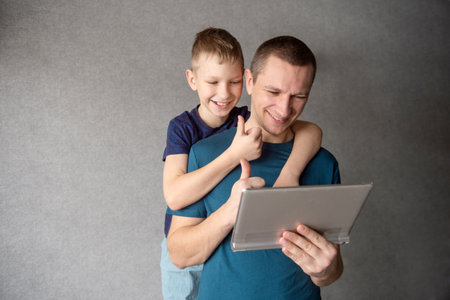 A cute boy hugs his dad from behind, talking on a video link in a tablet, showing a classの写真素材
