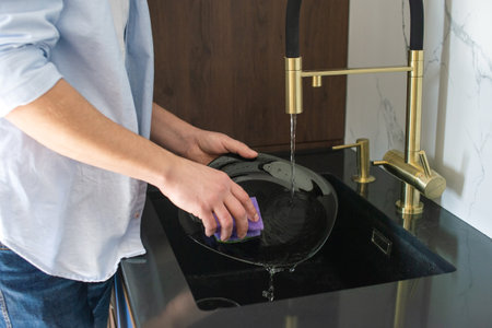 A man washes a black plate in the kitchen in the sink under water.の写真素材