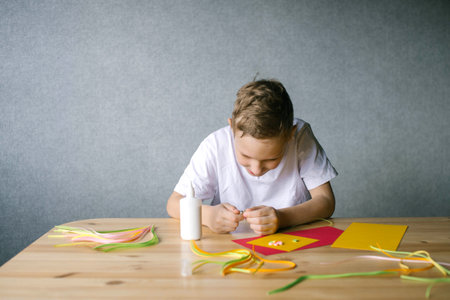 Cute boy makes details for a postcard by quilling method, sitting at a tableの写真素材