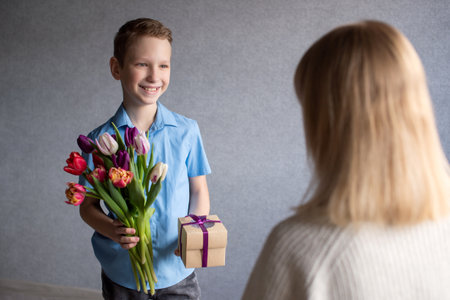 Cute boy smiles and gives mom flowers and a craft box with a birthday bowの写真素材