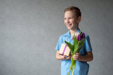 A cute boy is holding a colorful bouquet of tulips and a box with a gift in his hands.