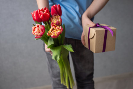 The boy hides behind his back a bouquet of red tulips and a box with a gift for momの写真素材