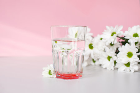A glass glass with water stands on a pink background with white chrysanthemum flowers in the morningの写真素材