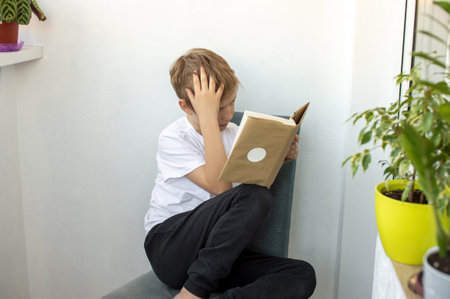 A tired boy sits on a chair with a book and holds his head in his hand. Reading a book sitting on...