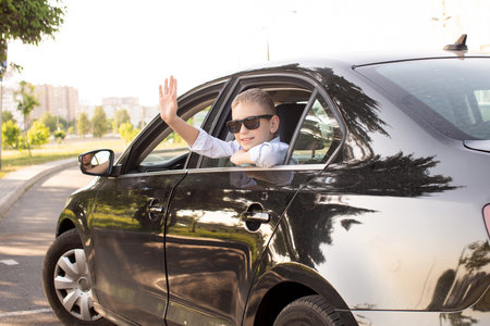 A cute boy climbed out of the car window wearing sunglasses and laughing. Waving to friends. contour light. Saying goodbye before leaving, on the move, going on vacationの写真素材