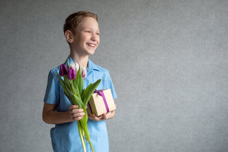 A cute boy laughs and holds a colorful bouquet of tulips and a box with a gift in his hands.