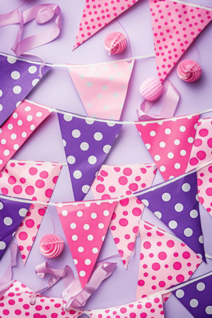 Purple and pink paper flags with polka dots and pom-poms on a pastel pink background. Party decoration concept with copy space for design and printの素材