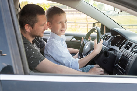 Dad and son in the front seat of a car driving a black car. The boy holds the steering wheel. Father...