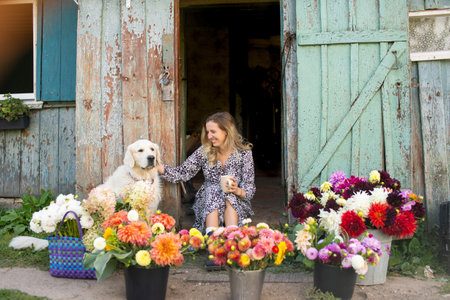 Cheerful woman sitting with a dog surrounded by vibrant flower baskets outside a rustic houseの写真素材