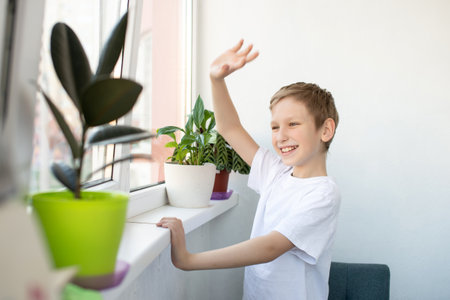 A cute boy looks out the window of a white balcony, waves to his friends and laughs. The boy is standing near the window with flowersの写真素材