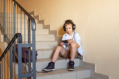 A cute schoolboy is sitting on the stairs with headphones and looking at the phone after school. Spend time on social networks. Throw the briefcase on the landing. Social problems of adolescentsの写真素材