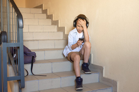 A frustrated boy is sitting on the stairs with a phone in his hands and headphones, holding his head, worried. Getting bad newsの写真素材