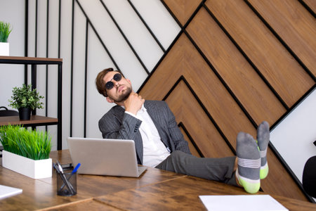A man is sitting in the office with a laptop, his feet are on the table. Procrastinating in sunglasses. Can't get to work. Spend time in the office.の写真素材
